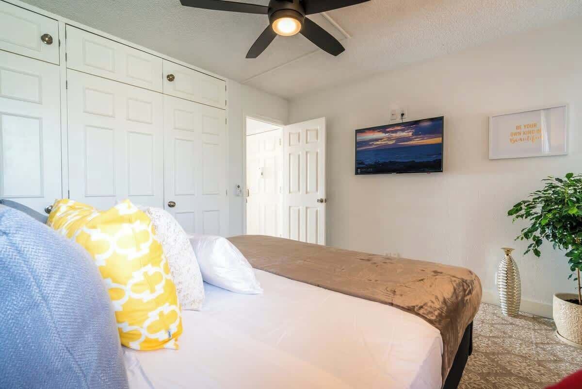 Un dormitorio con cama, armario blanco, ventilador de techo, cuadro enmarcado y planta en maceta.