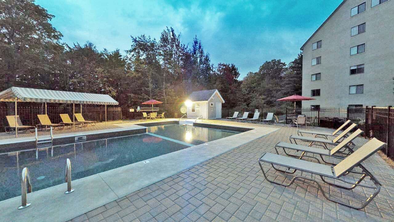 A swimming pool area with lounge chairs and umbrellas, a small building, and a multi-story building in the background.