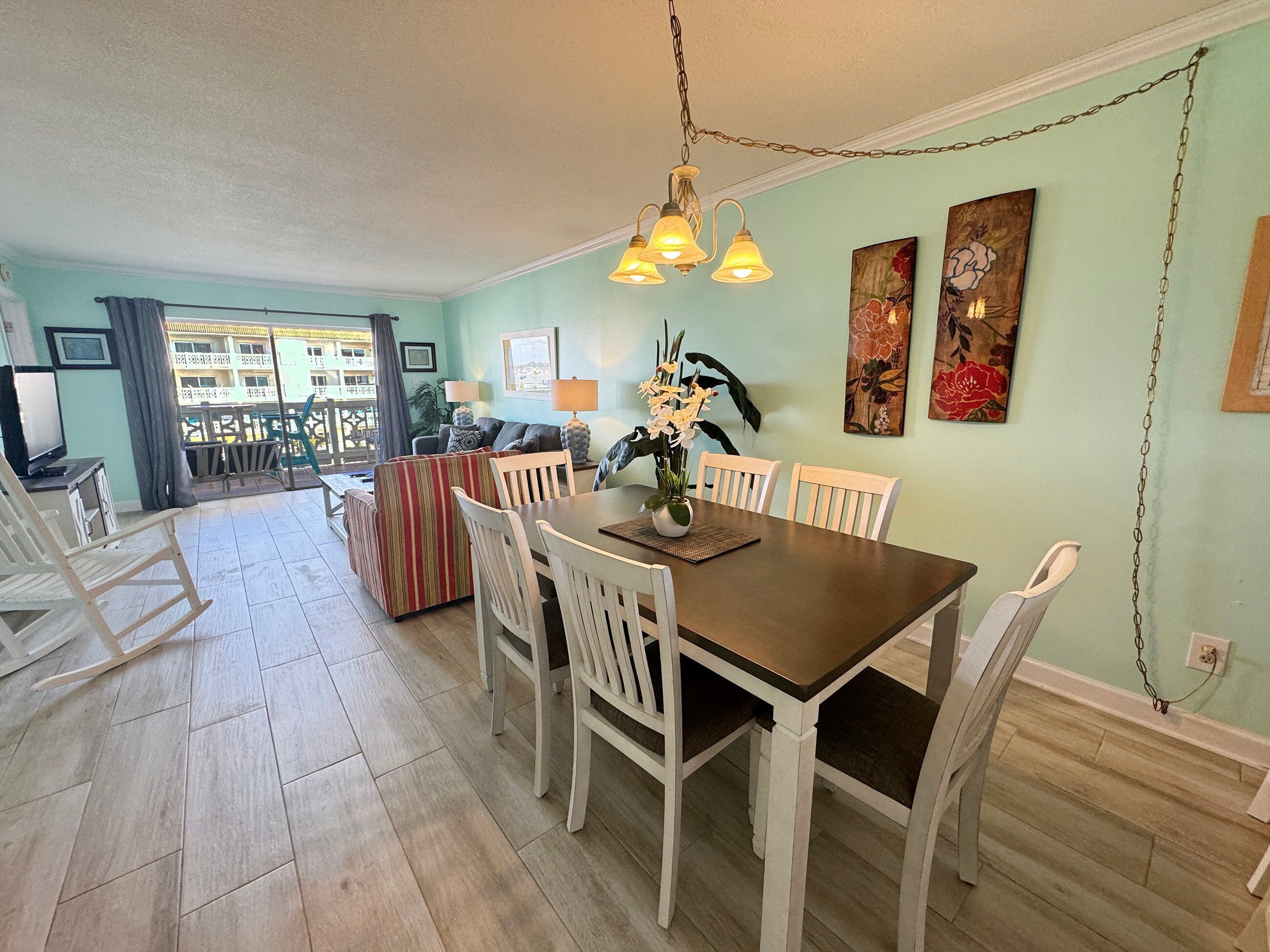 A dining area with a wooden table, white chairs, and a chandelier. There are two paintings on the wall and a potted plant on the table.