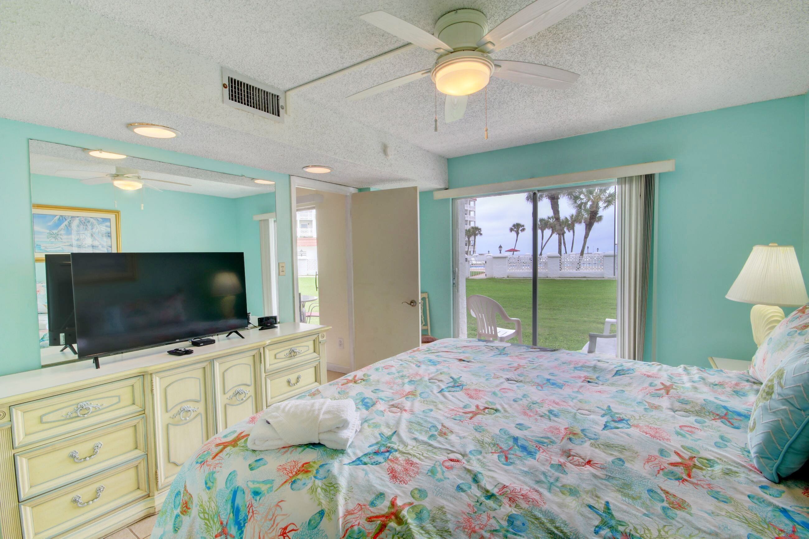 A bedroom with a bed, a television, a dresser, and a window with a view of palm trees.