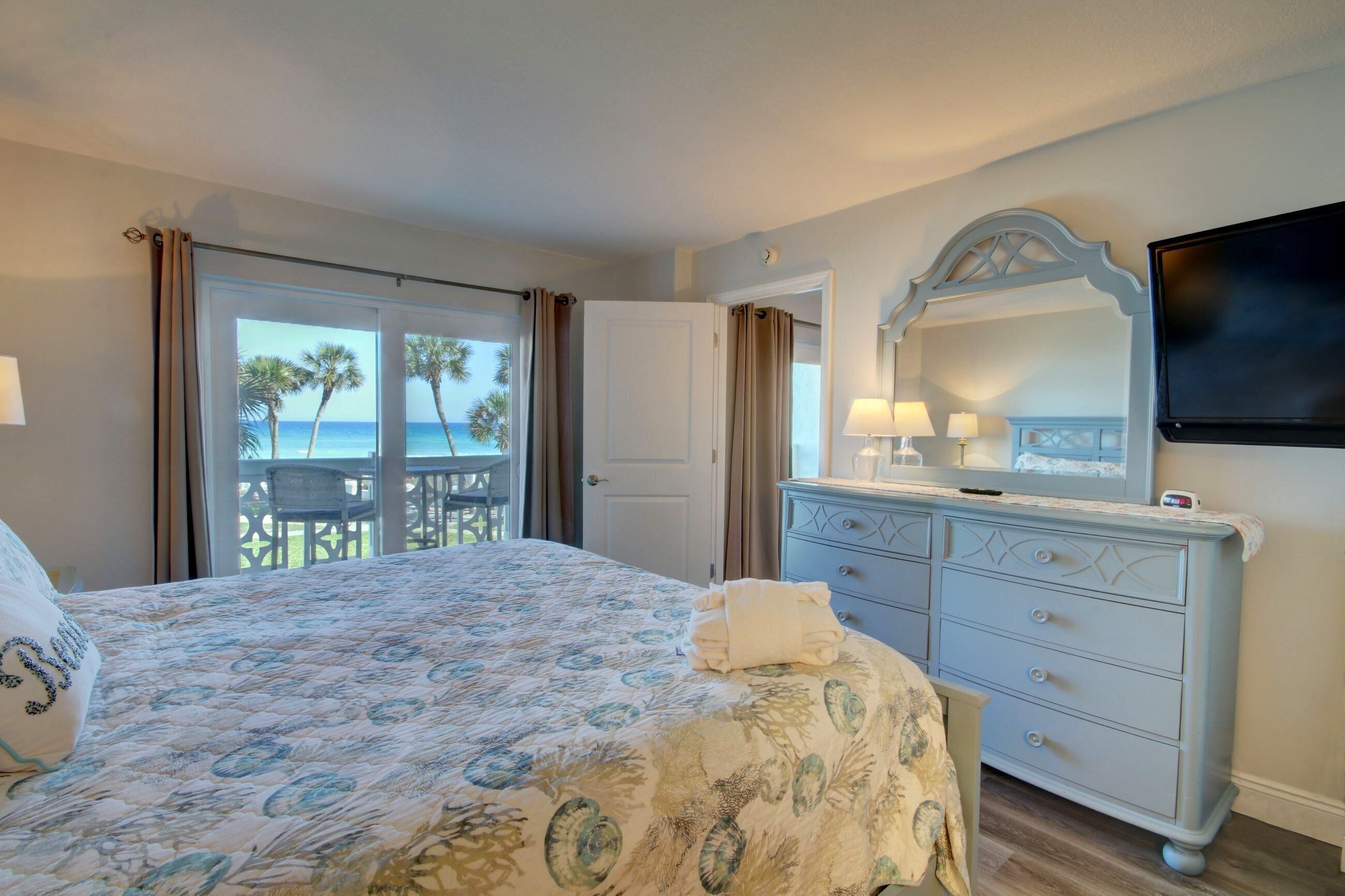 A bedroom with a bed, a dresser, a TV, and a balcony with a view of palm trees and the sea.