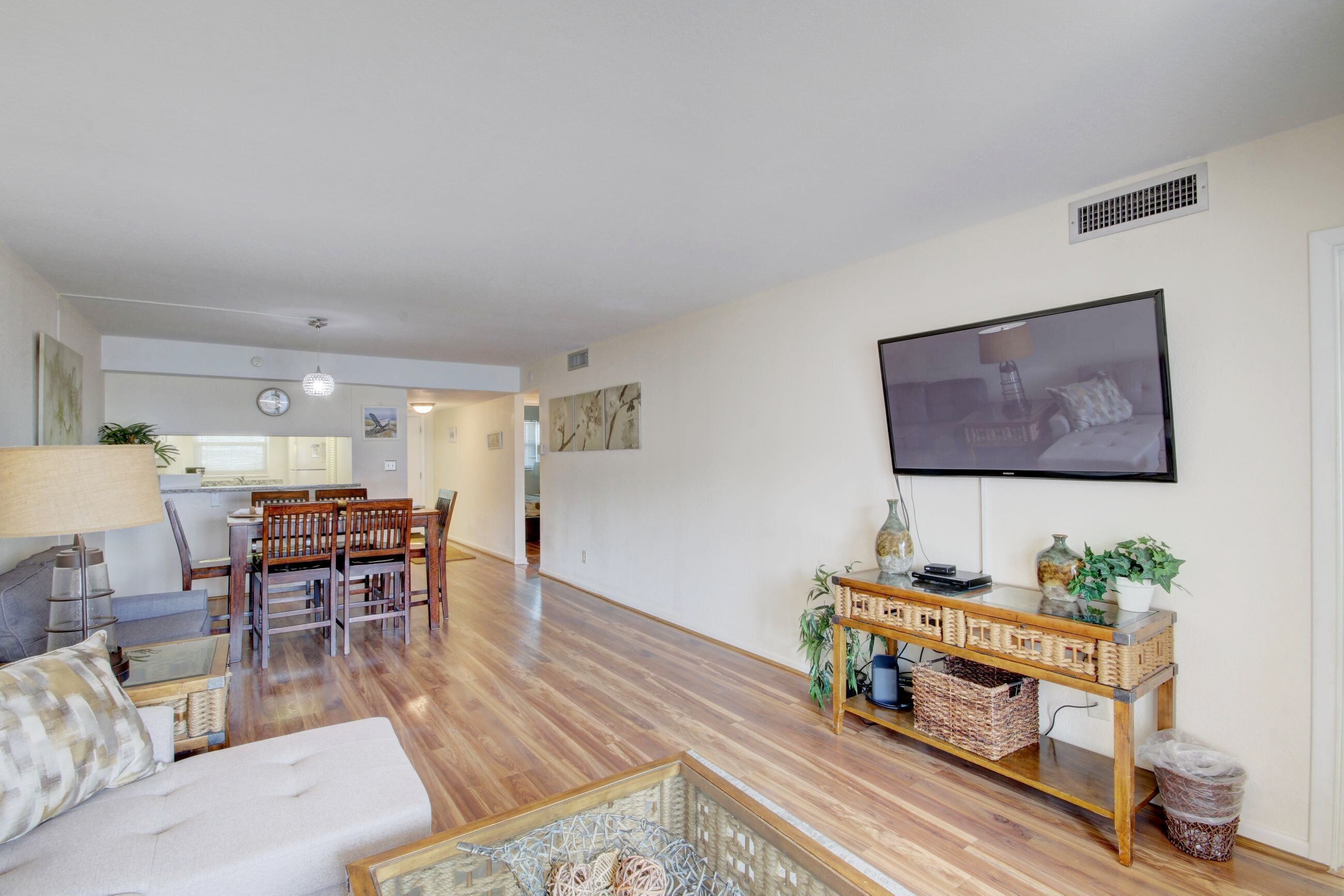 A living room with a sofa, dining table, chairs, a TV mounted on the wall, and a small table with decorative items.