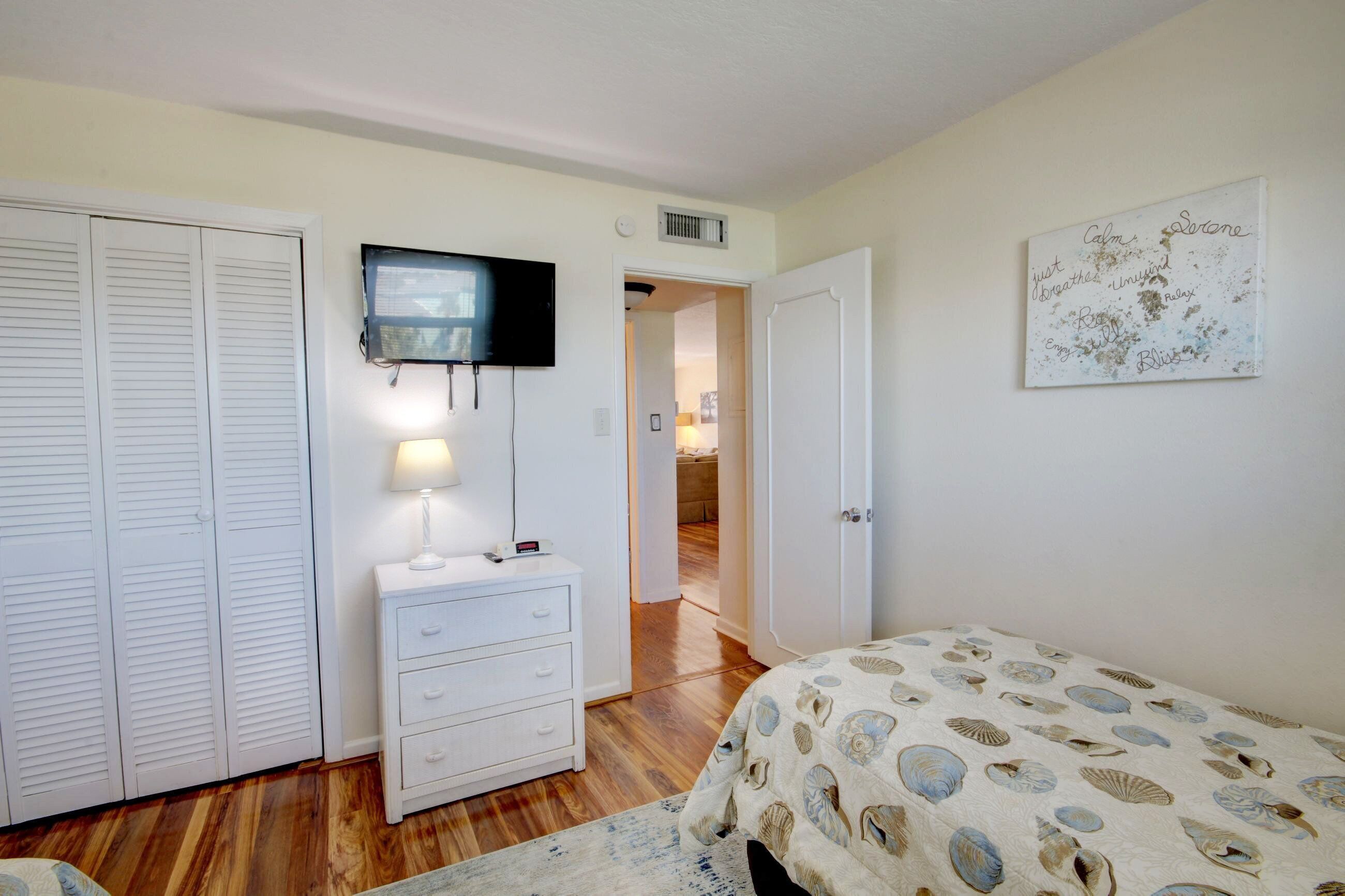 Una camera da letto con un letto, una TV a parete, un comodino con una lampada e una porta che dà su un'altra stanza.