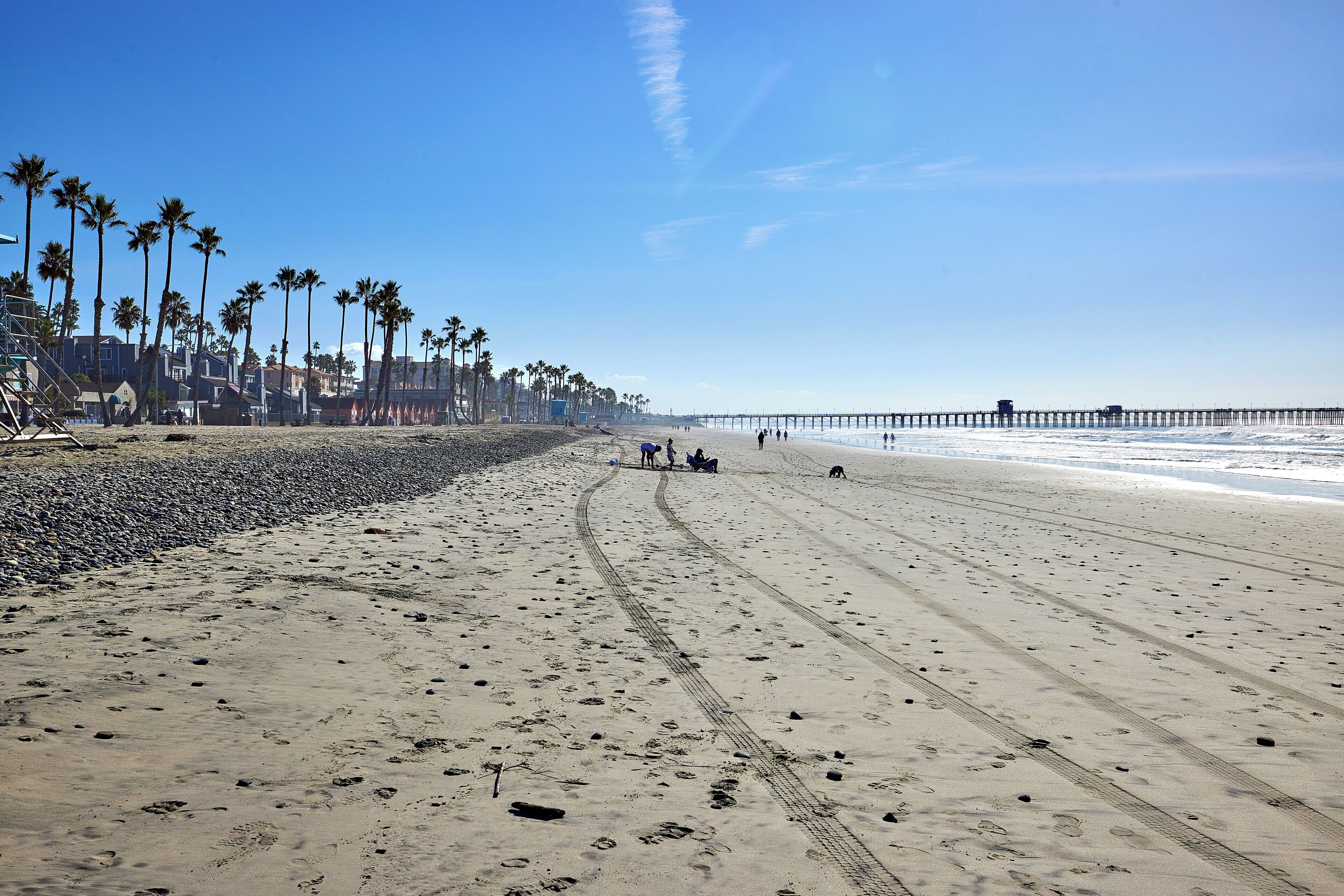 A beach with a long pier extending into the ocean, palm trees lining the shore, and footprints in the sand.