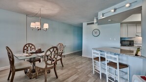 Un comedor con mesa de vidrio y sillas de madera, una araña, y una cocina con armarios blancos y un microondas.