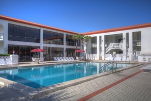 Une piscine entourée de chaises longues et de parasols, bordée d’un bâtiment doté de grandes fenêtres et d’un toit rouge.