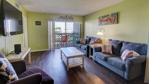 Un salón con sofá, una mesa de centro y televisor. Hay un balcón con vistas a edificios y un letrero que dice 'family'.