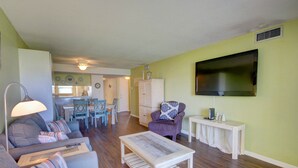 Una sala de estar con un sofá, sillón, mesa de centro y televisor. Se observa un comedor con una mesa y sillas al fondo.