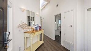 Um corredor com piso de madeira, uma porta branca, um console com ramos decorativos e um claraboia.