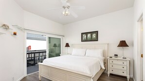 Quarto com cama grande, mesa de cabeceira, ventoinha no teto e porta de vidro para uma varanda.