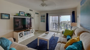 A living room with a TV, white entertainment unit, blue carpet, beige sofa, and a balcony with a view. - 141 El Matador (Fort Walton Beach)