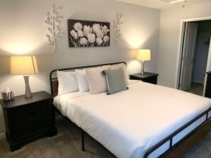 Een net opgemaakt bed met witte beddengoed, twee nachtkastjes met lampen, een bloemrijk schilderij aan de muur en een deur op de achtergrond.