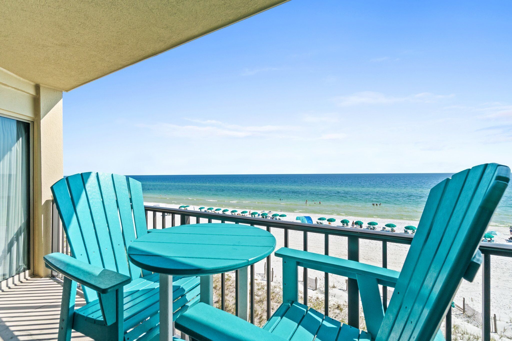 A balcony with turquoise Adirondack chairs and a table, overlooking a beach with umbrellas and people.