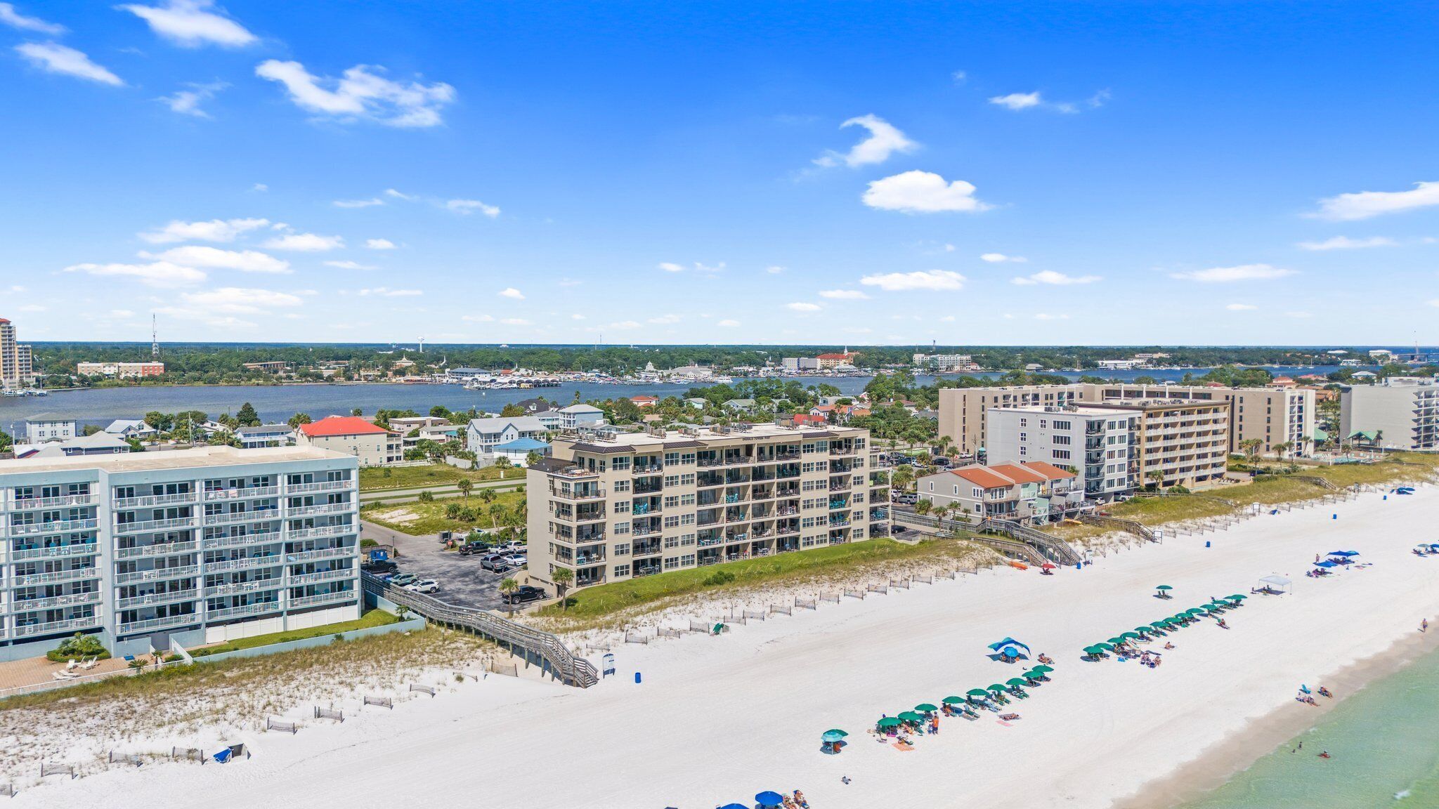 A coastal hotel with multiple floors, a beach with umbrellas, and a view of the sea and cityscape.