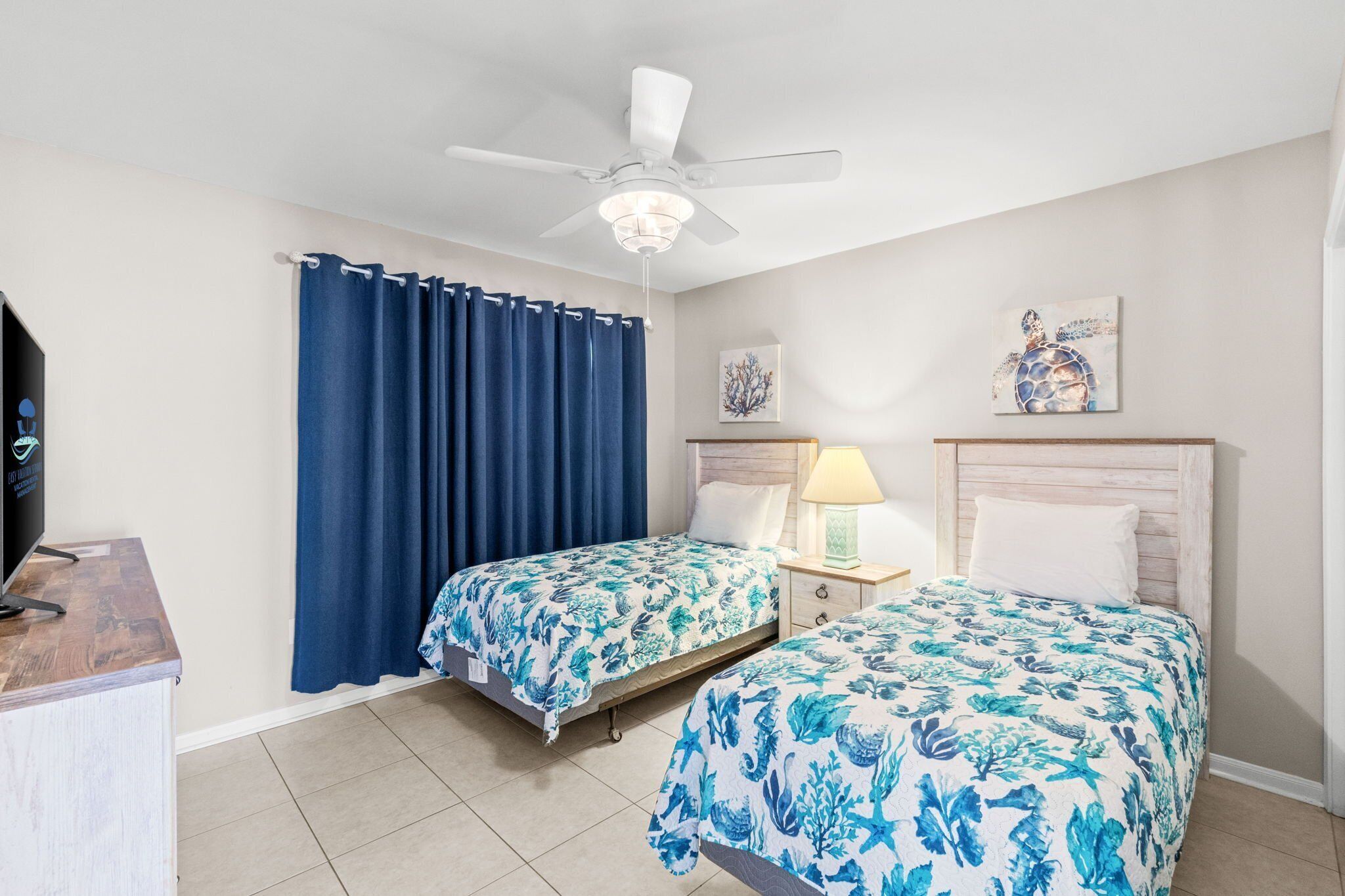 A hotel room with two beds, a ceiling fan, a TV, and artwork on the walls.