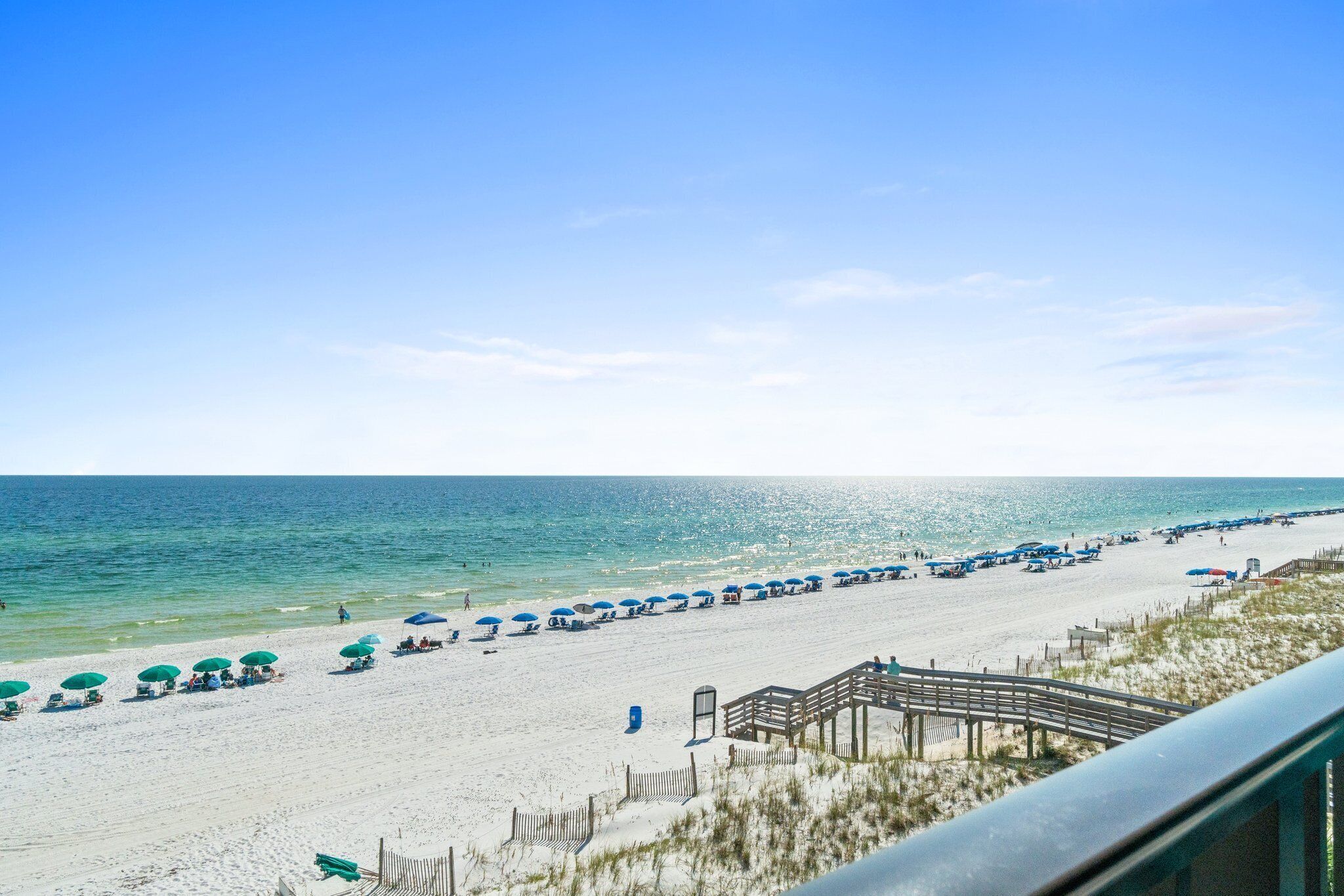 Ein Strand mit Reihen blauer Sonnenschirme und Liegestühle, eine Holzpromenade und das Meer im Hintergrund.