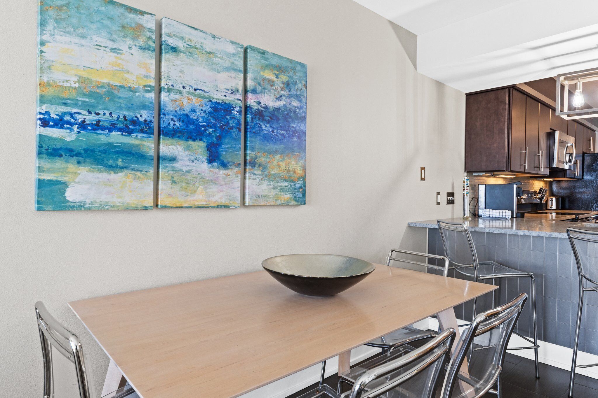 A modern kitchen with a central island, stainless steel appliances, and a dining area with a wooden table and clear chairs.