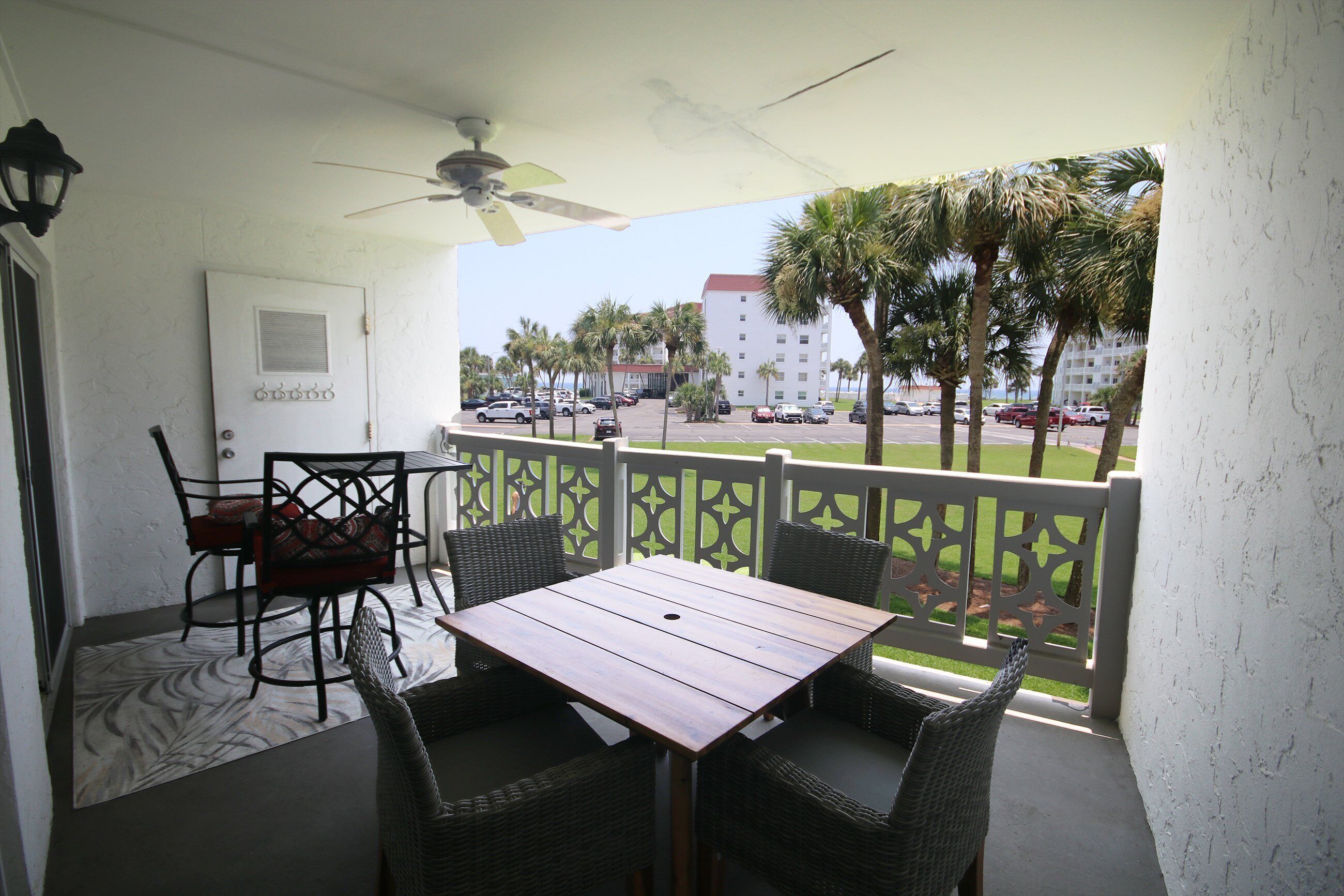 A balcony with a table and chairs, a ceiling fan, and a view of a parking lot and palm trees.