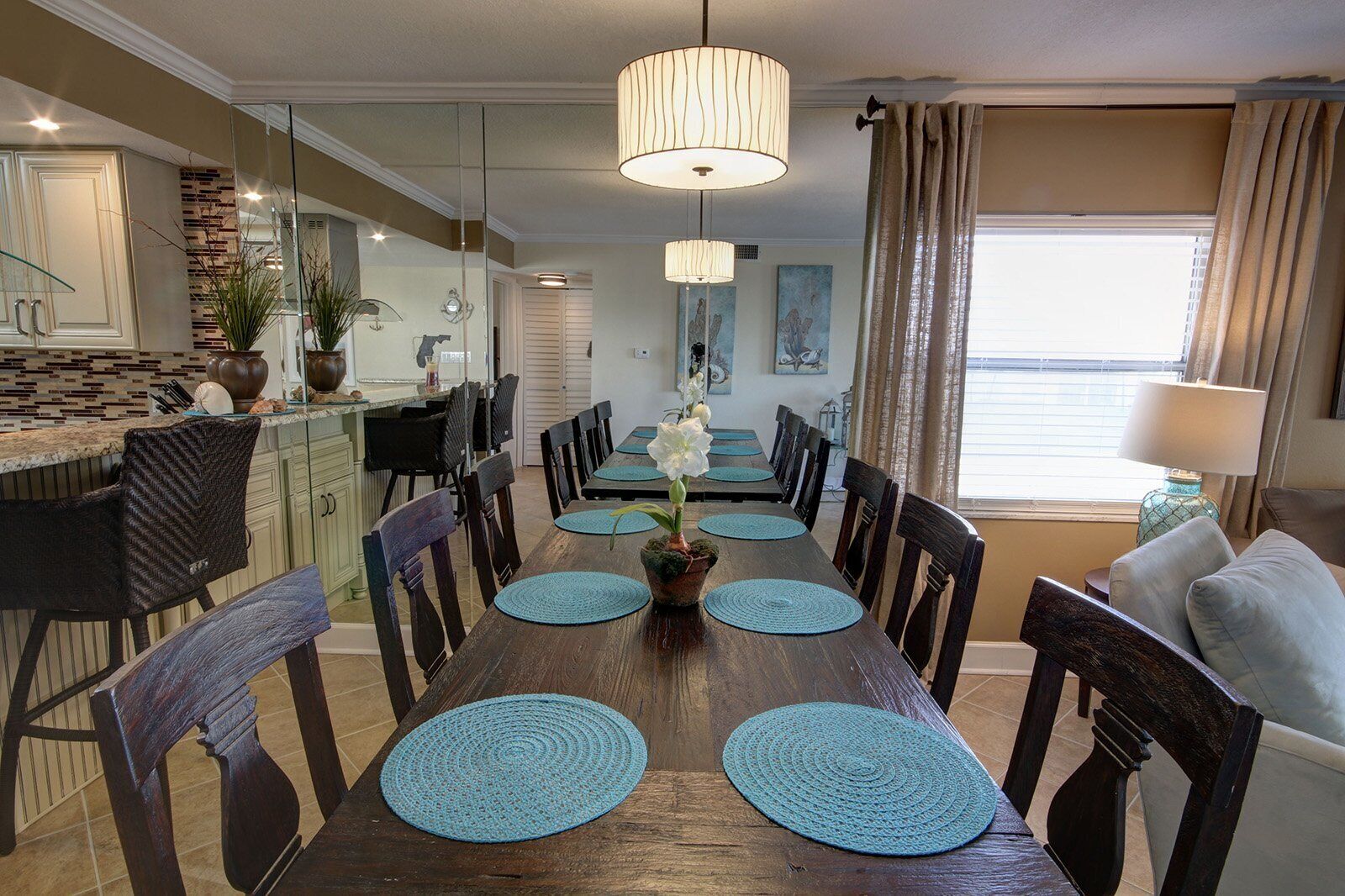 Een eetgedeelte met een lange houten tafel, blauwe placemats en een centraal bloemstuk. Er staan stoelen rond de tafel en links is een keuken te zien.