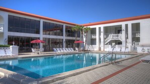 Ein Poolbereich mit Liegestühlen und Sonnenschirmen, ein Gebäude mit großen Fenstern und einem roten Dach, sowie ein klarer, blauer Himmel.