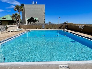 A clear blue pool with a 'NO DIVING' sign, lounge chairs, and a building with a green roof in the background. - Emerald Isle 102 - The Seashell (Fort Walton Beach)