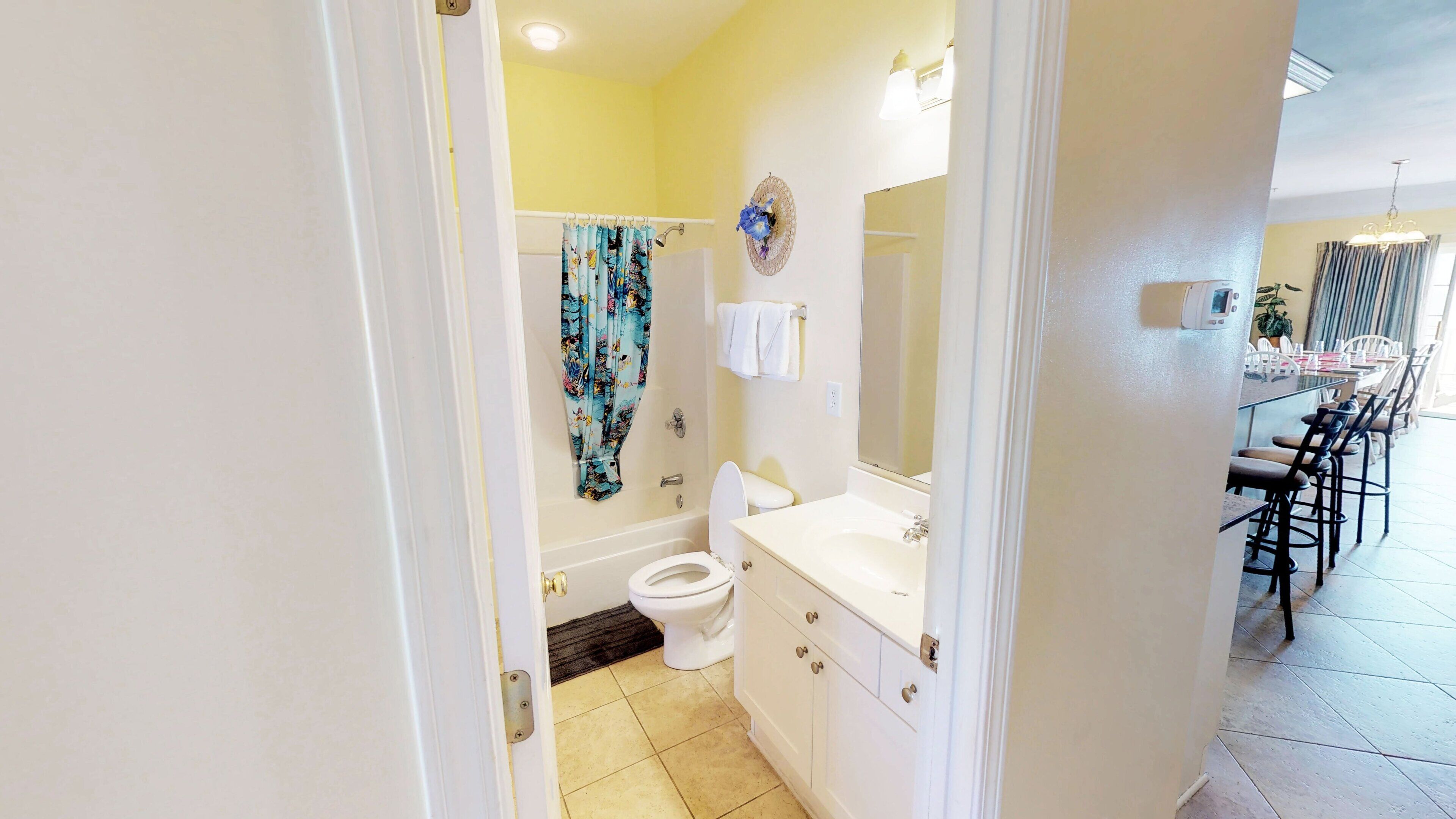 A bathroom with a toilet, sink, and shower. There is a dining area visible in the background.