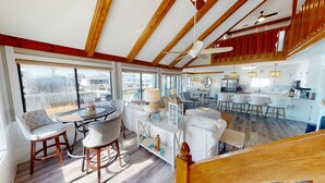 A spacious living area with a high ceiling, large windows, and a view of the outside. There is a dining area with a round table and chairs, and a kitchen with modern appliances. - Wait & Sea (North Myrtle Beach)