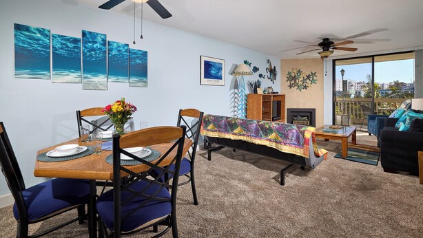 A living room with a dining area, ceiling fans, artwork, and a view through large windows. - North Coast Village B212 (Oceanside)