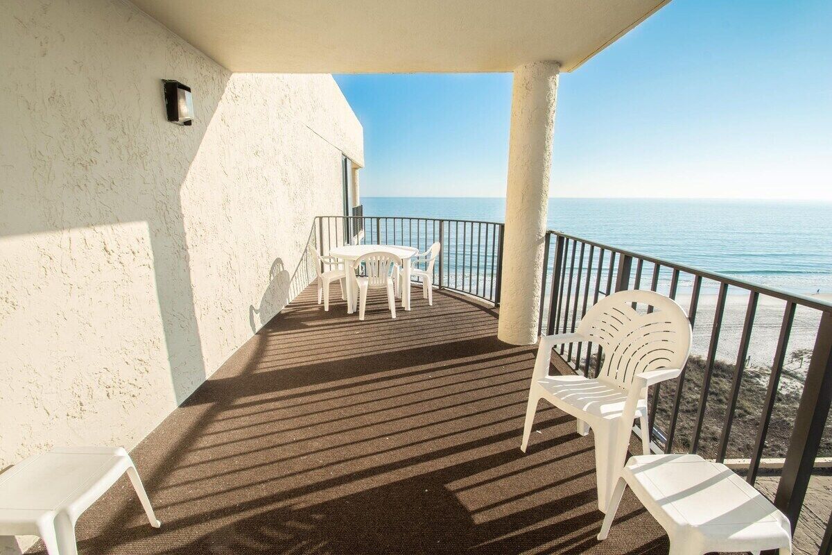 A balcony with a view of the sea, featuring a table and chairs, a railing, and a textured wall.