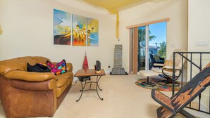 A living room with a brown leather sofa, a small table, and a chair. There are abstract paintings on the wall and a view of the outside through a glass door. - Oceanview Beach Home - 209w (Oceanside)