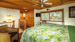 Une pièce avec un plafond en bois, un ventilateur de plafond, un lit recouvert d’une courtepointe à motifs tropicaux, une chaise en bois, une petite table, un téléviseur et une fenêtre avec des stores.