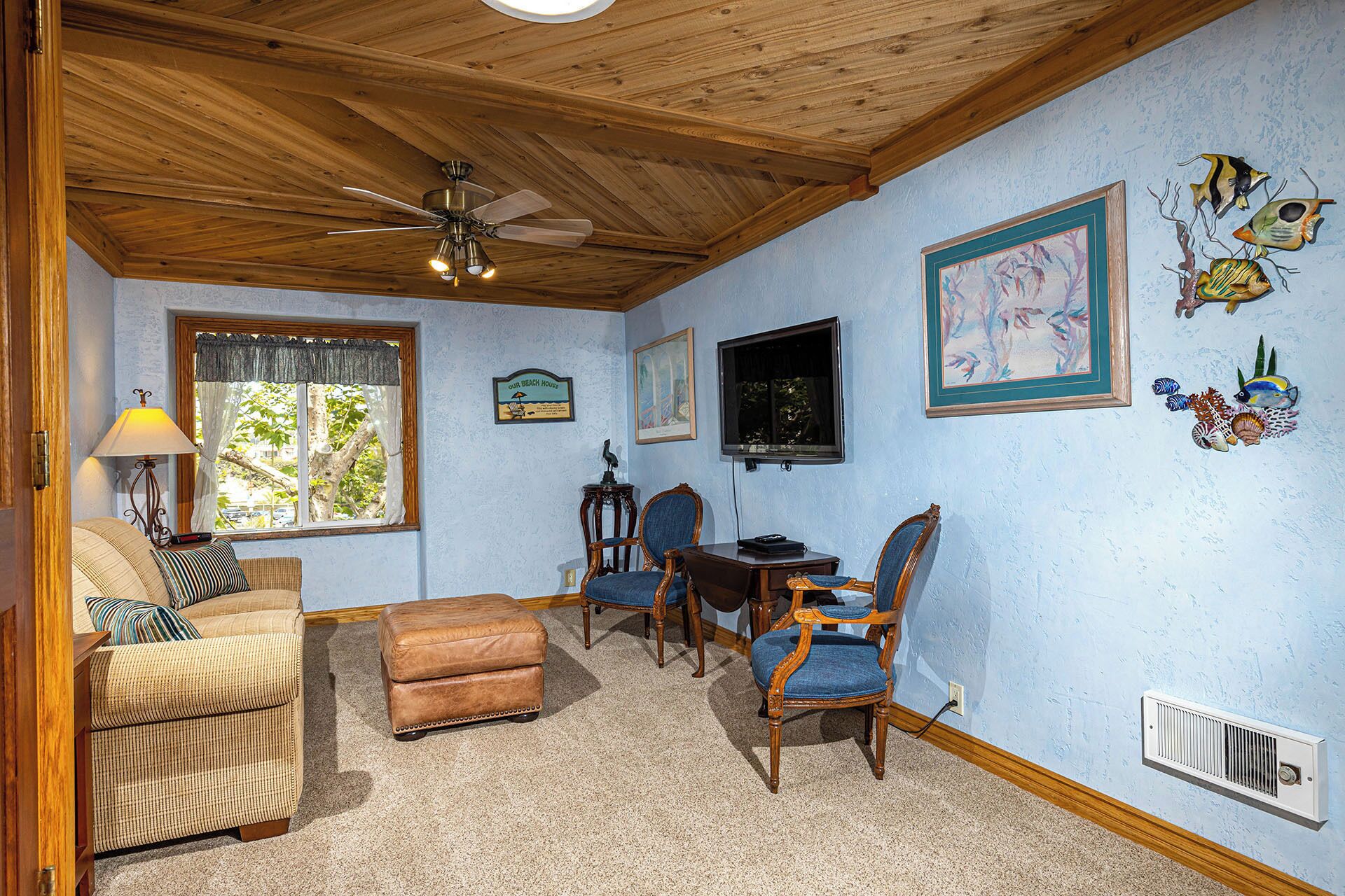 A cozy room with a wooden ceiling, a ceiling fan, a window with curtains, a dining table with chairs, and wall decorations including framed art and fish.