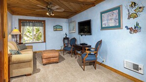 Une pièce chaleureuse avec un plafond en bois, un ventilateur de plafond, une fenêtre avec des rideaux, une table à manger avec des chaises et des décorations murales comprenant des œuvres d’art encadrées et des motifs de poissons.