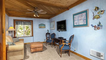 A cozy room with a wooden ceiling, a ceiling fan, a window with curtains, a dining table with chairs, and wall decorations including framed art and fish.