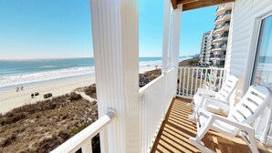 Un balcón con sillas de plástico blanco, una terraza de madera y vista a la playa y al océano.