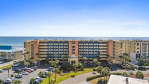 A multi-story hotel with a central entrance, parking lot, and a view of the ocean. - Pelican Isle 113 - Salty Seahorse (Fort Walton Beach)
