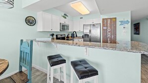 A kitchen with white cabinets, a stainless steel refrigerator, a microwave, and a sink. There are two black stools at the island. - Pelican Isle 113 - Salty Seahorse (Fort Walton Beach)