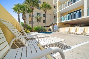 A poolside area with white lounge chairs, a pool, palm trees, and a multi-story building with balconies. - Pelican Isle 208 - Sandy Shores (Fort Walton Beach)