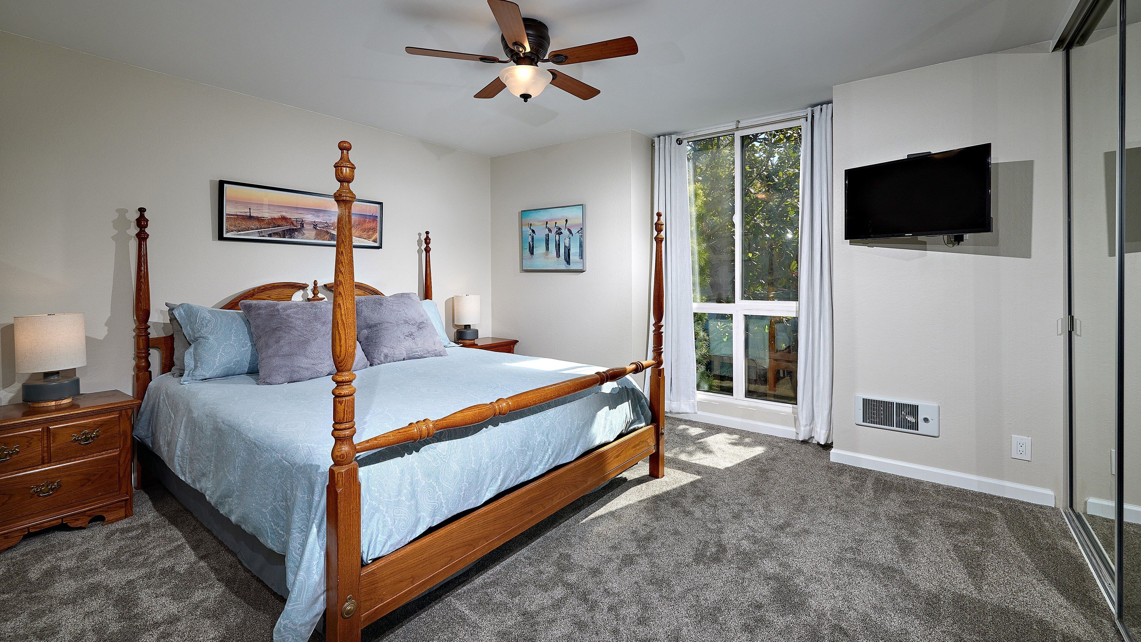 A bedroom with a four-poster bed, a ceiling fan, a large window with curtains, a flat-screen TV, and a nightstand with a lamp.