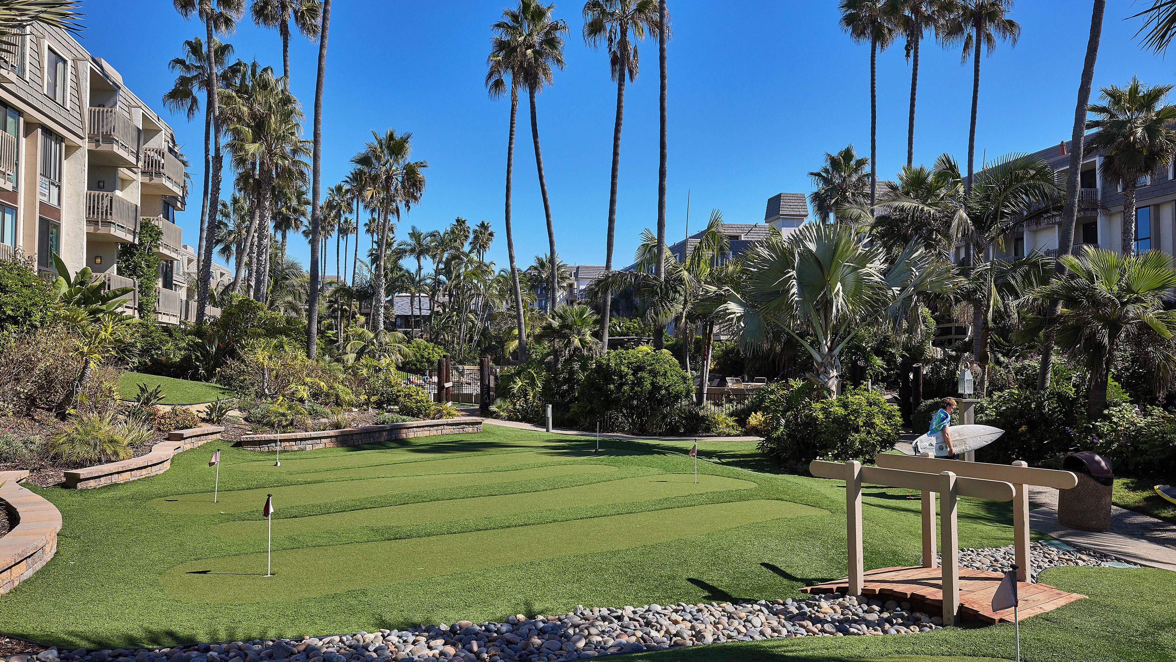 A well-maintained golf course with a putting green, surrounded by palm trees and a stone wall.