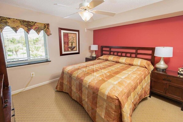 A bedroom with a bed, a nightstand, a lamp, a ceiling fan, a window with blinds, and a wall with a floral curtain. - Yacht Club 2-805 (North Myrtle Beach)