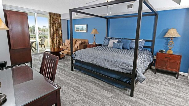 A four-poster bed with a blue and gray patterned bedspread, a wooden nightstand with a lamp, a wooden chair, and a dining table with chairs. - Yacht Club 2-704 (North Myrtle Beach)