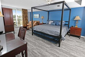 Ein Bett mit Baldachin, mit einer blau-grauen Musterdecke, ein Nachttisch aus Holz mit einer Lampe, ein Holzstuhl und ein Esstisch mit Stühlen.