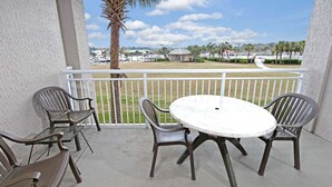 A covered outdoor area with a round table and four chairs, overlooking a grassy area with palm trees and a distant view of buildings. - Yacht Club 2-704 (North Myrtle Beach)