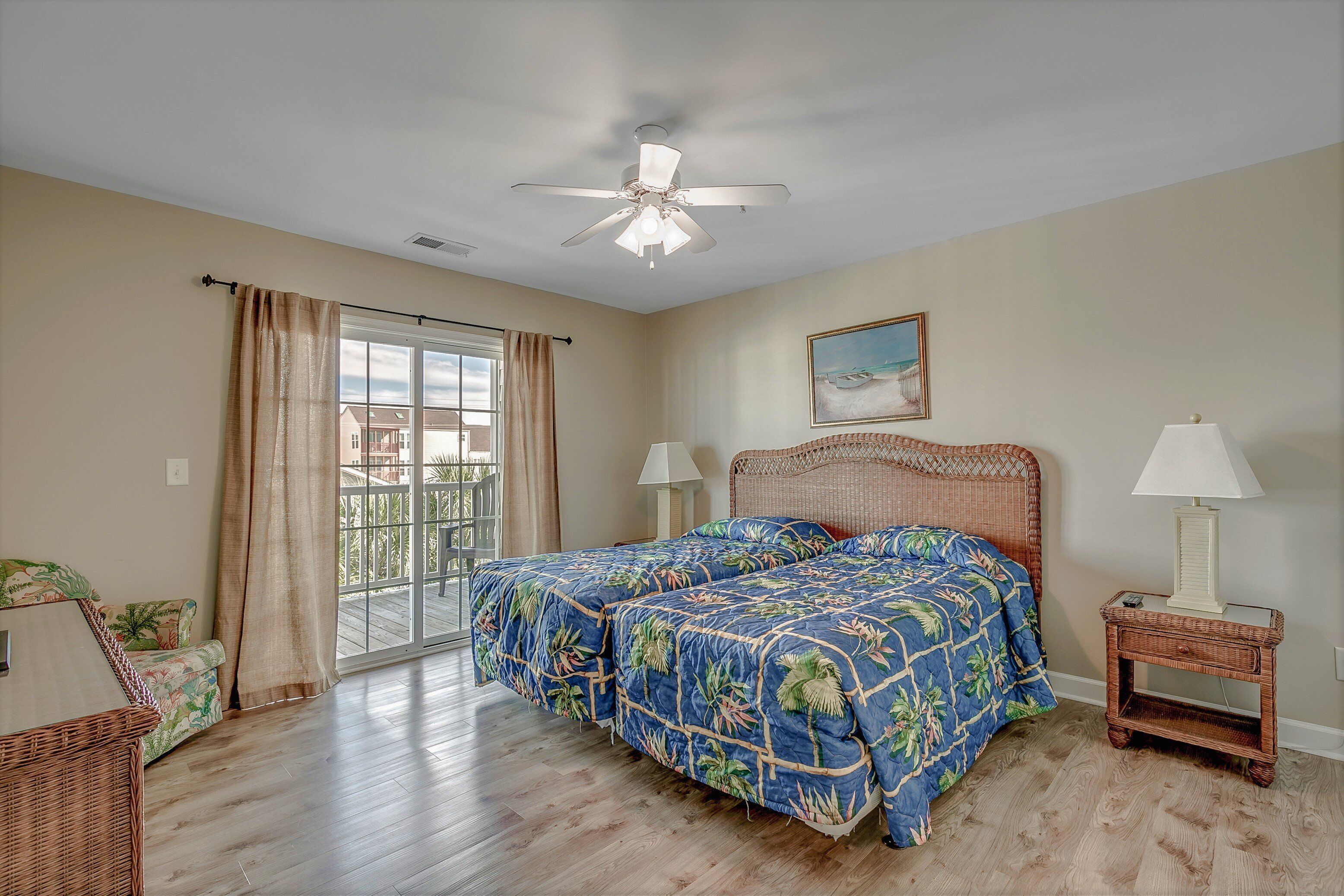 A bedroom with a bed, two bedside tables with lamps, a ceiling fan, a large window with curtains, and a view of the outdoors.