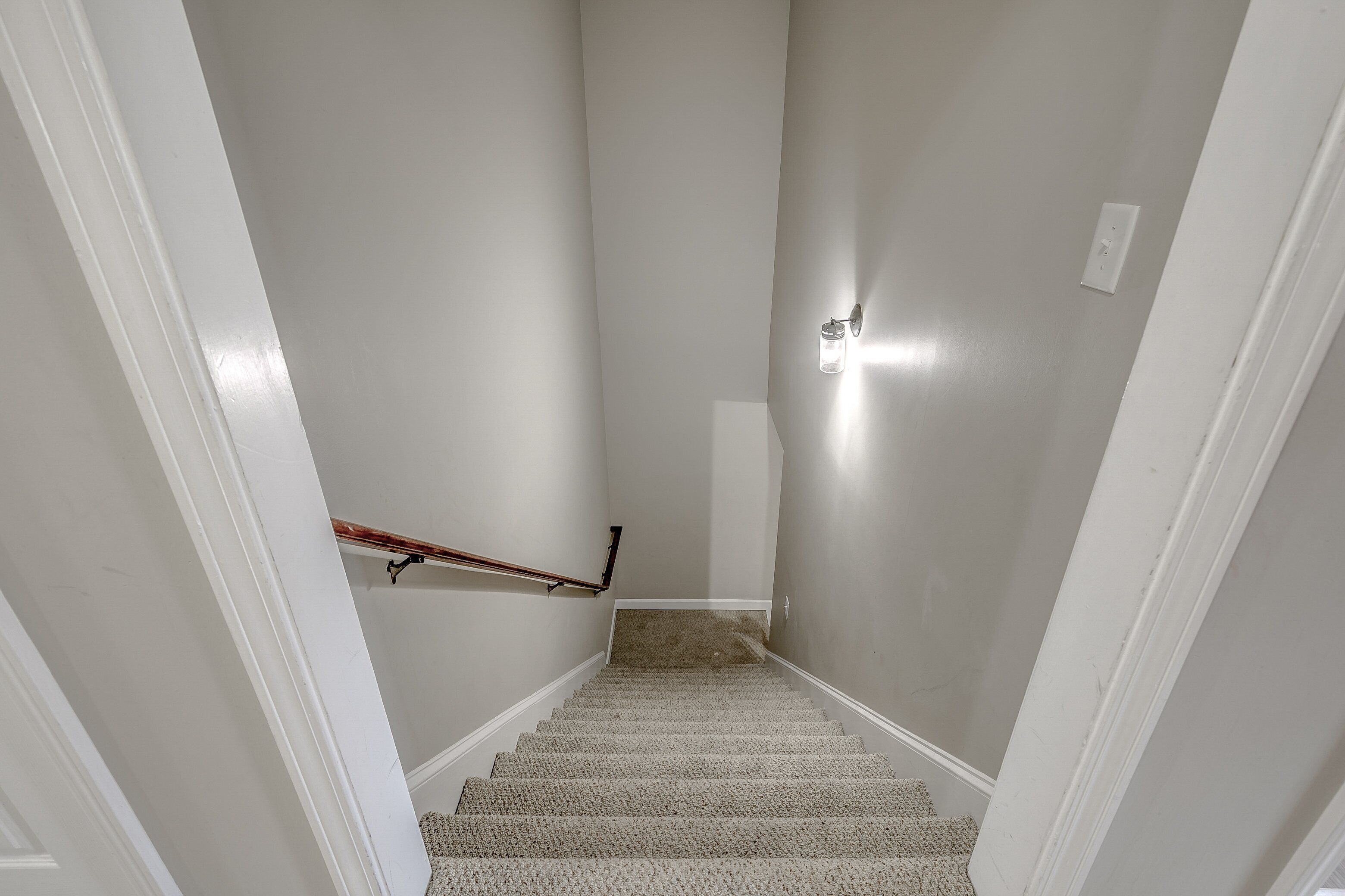 Una escalera alfombrada con barandilla, paredes blancas y un interruptor de luz en la pared.