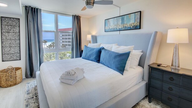 Habitación de hotel con una cama grande, un ventilador de techo, vista al océano, un cuadro en la pared y una mesita de noche con una lámpara.