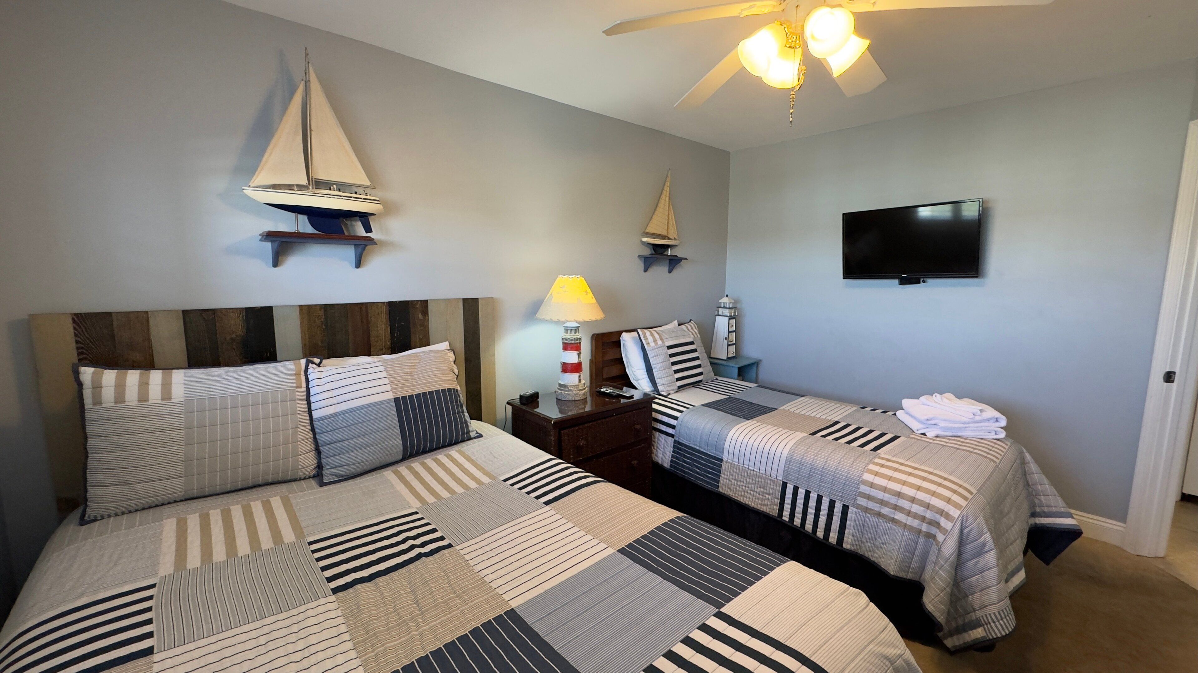 A bedroom with two beds, a ceiling fan, a TV, and decorative sailboat models.