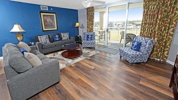 A living room with a blue wall, a ceiling fan, a sofa, a patterned armchair, and a view of the outdoors through a sliding glass door. - Yacht Club 2-404 (North Myrtle Beach)