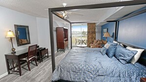 A four-poster bed with a blue patterned bedspread, a desk with a lamp, a chair, and a view of the outdoors through a balcony. - Yacht Club 2-404 (North Myrtle Beach)